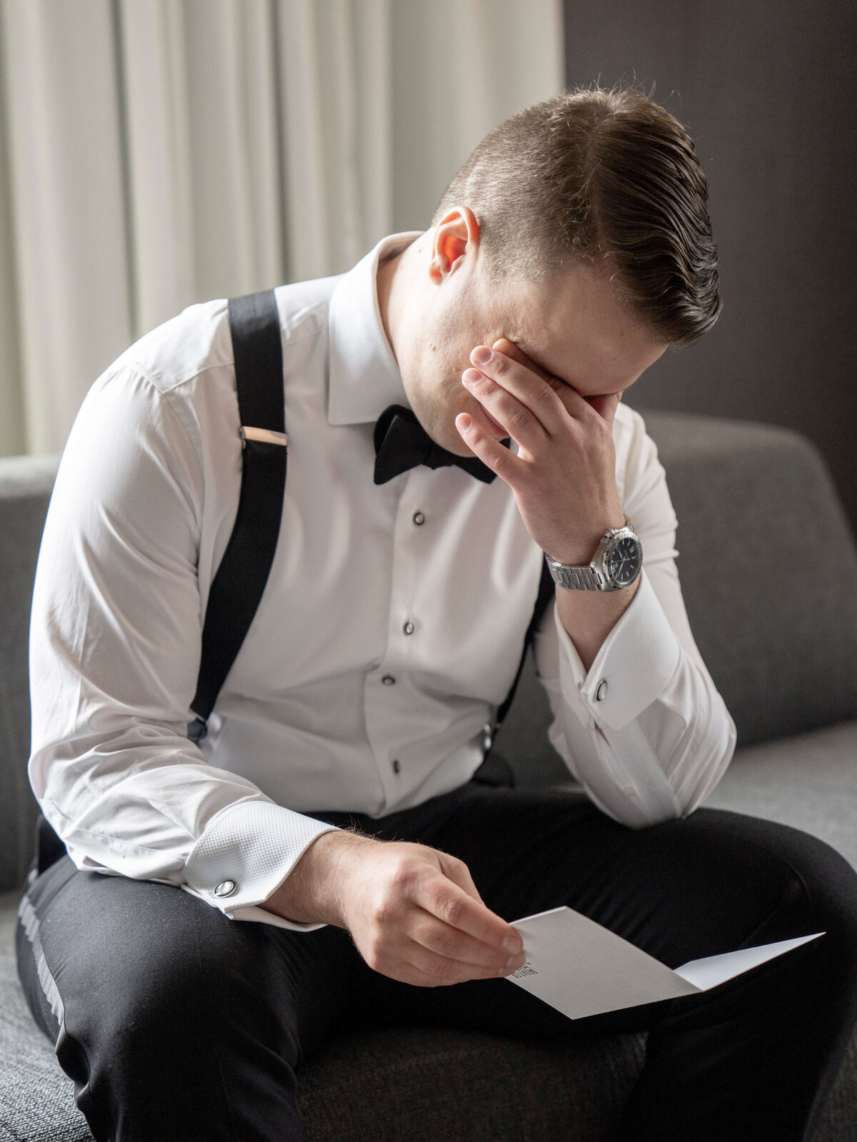 A man in formal attire sits on a couch, holding a letter and covering his face with one hand. He appears emotional.