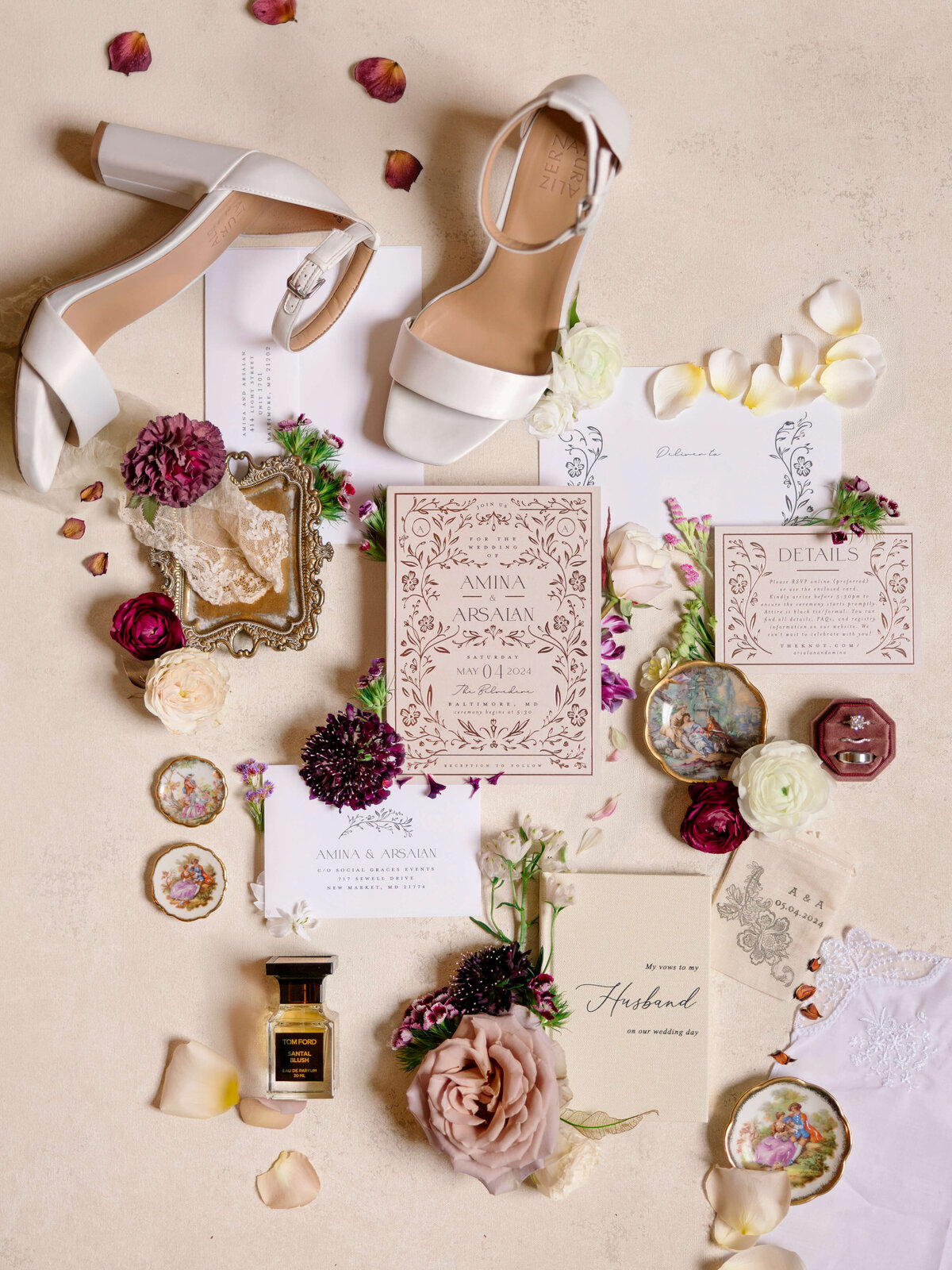 Flat lay of a wedding invitation suite surrounded by white heels, floral accents, perfume, and decorative dishes. The invitations have a floral design, and petals are scattered around.
