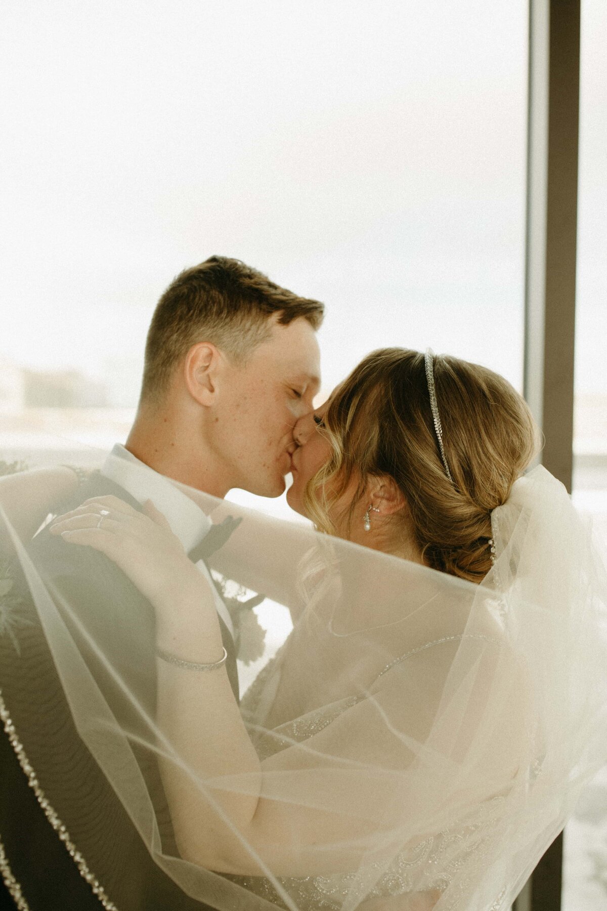 A bride and groom sharing a romantic kiss, with the bride's veil flowing gracefully around them, indoors at a winery with soft natural light illuminating the scene.