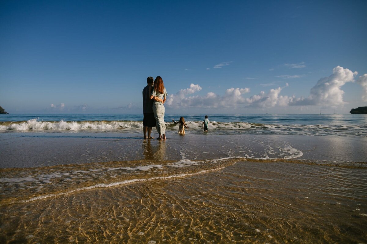 A husband and wife hold each other on the beach as their children run into the waves of the ocean.