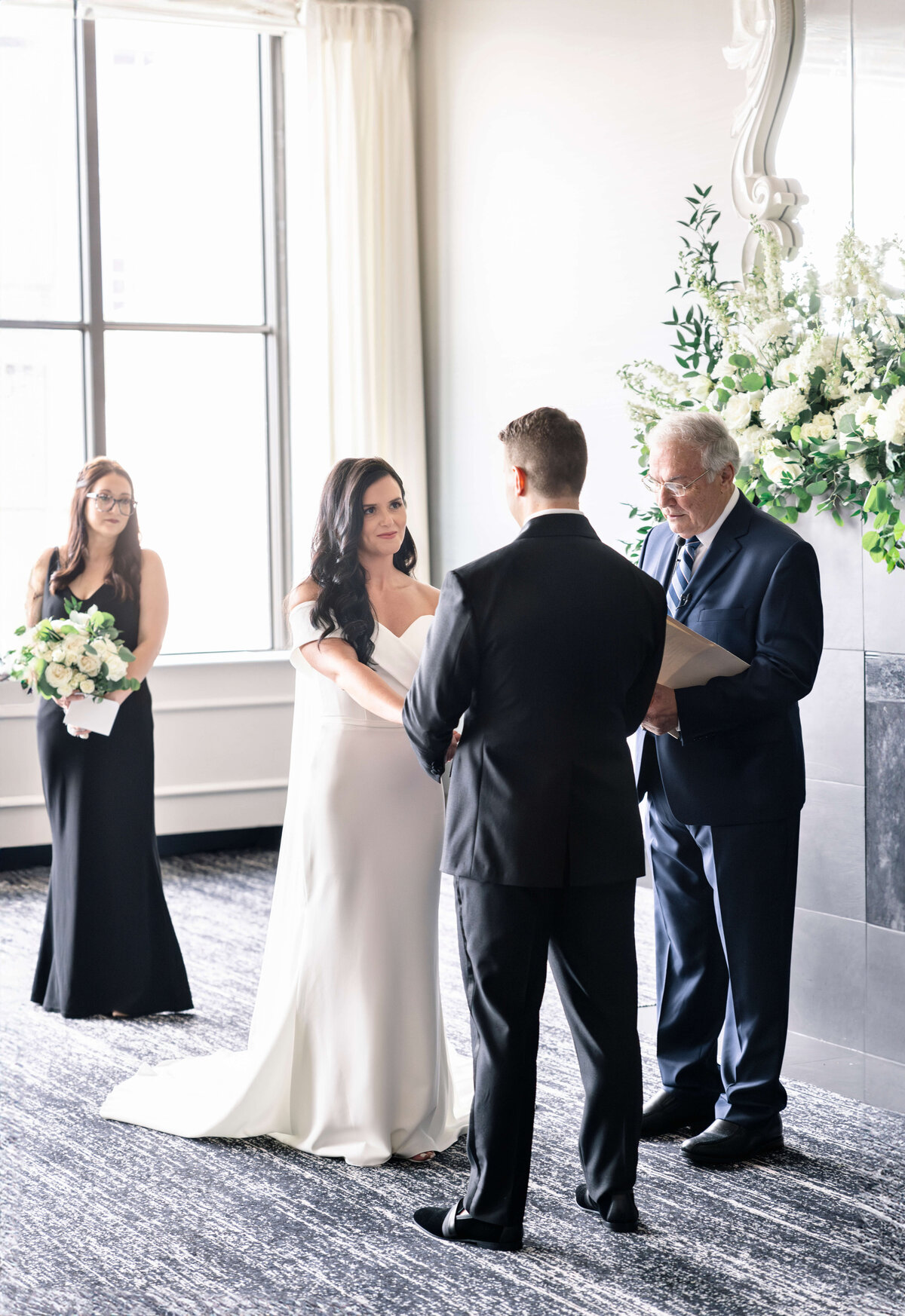 A bride and groom stand facing each other during a wedding ceremony. A man in a suit officiates with a paper in hand. A bridesmaid in a black dress holds a bouquet. White floral arrangements decorate the room.