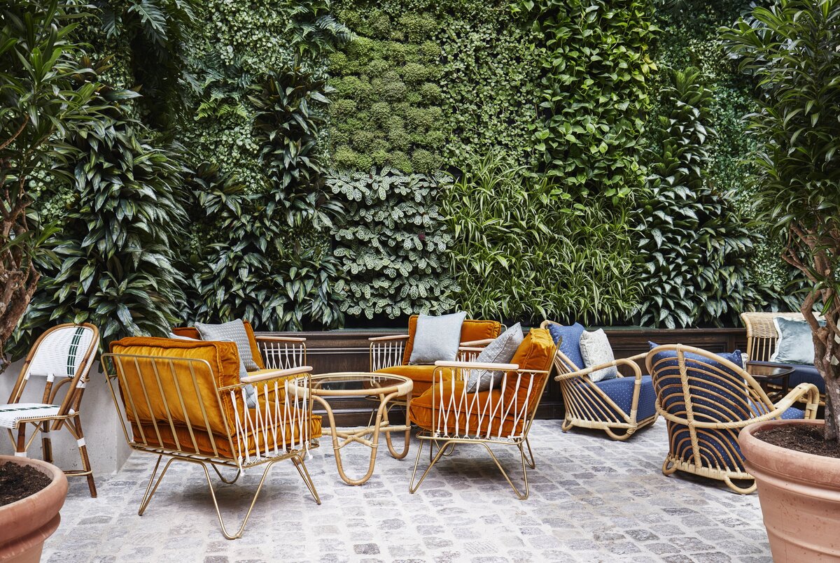 Outdoor area with cobblestone floor, green plant wall and brass lounge chairs with orange velvet cushions and mid century bamboo coffee table