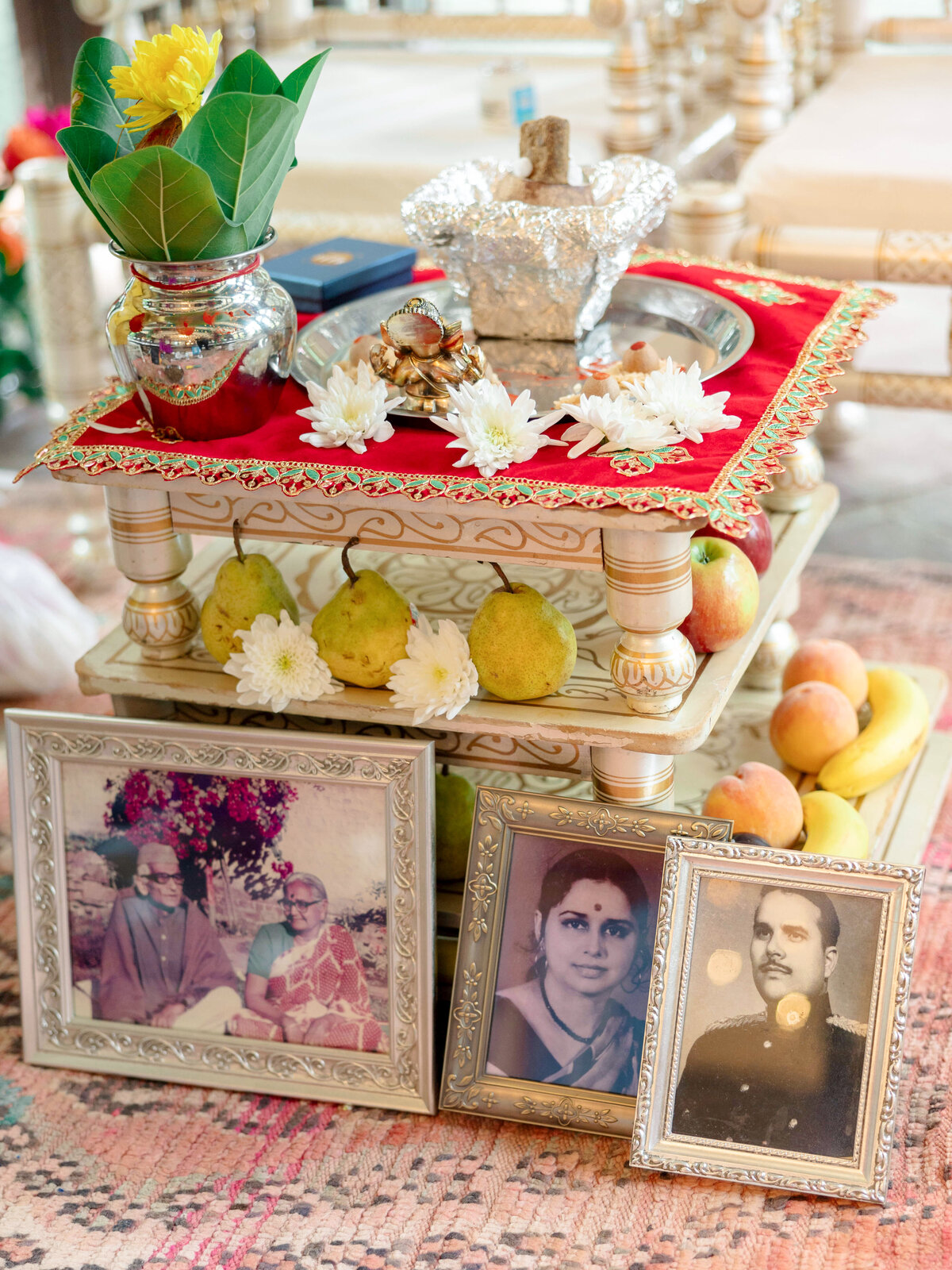 A decorative altar with framed photos of three individuals, adorned with flowers, leaves, and a silver-covered item. Pears and other fruits are placed around it. The altar is set on a patterned rug.