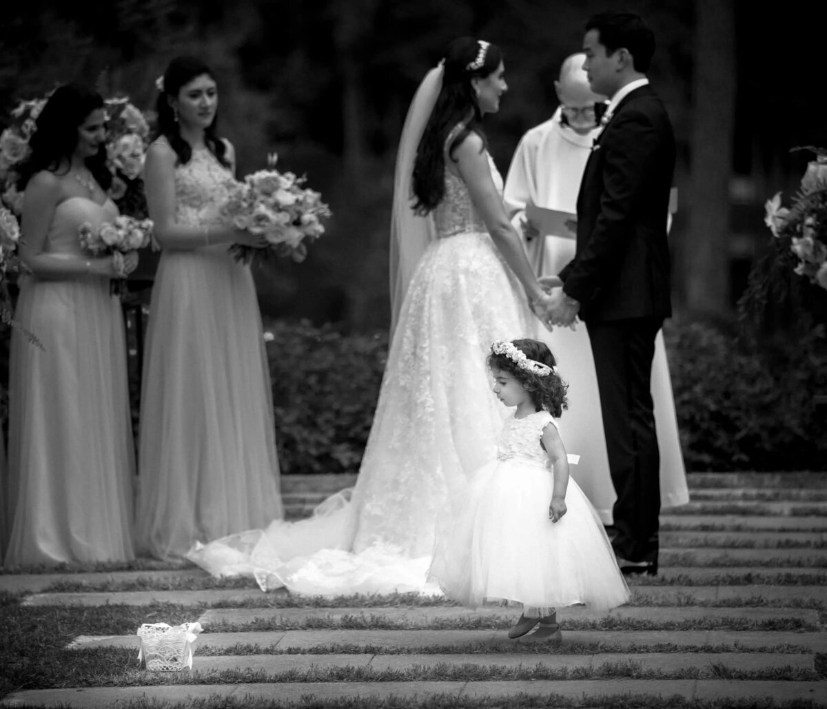 A flower girl in a fluffy dress looks up at the bride during a wedding ceremony, with the bridal party in the background in soft focus.