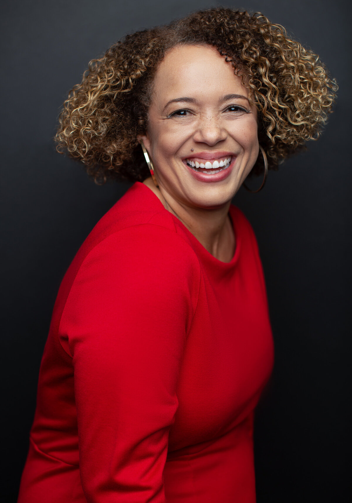 Cincinnati portrait showcasing a light-skinned woman with vibrant curly hair, beaming in a striking red top against a dark background. Her genuine smile and confident demeanor highlight the essence of modern studio photography.