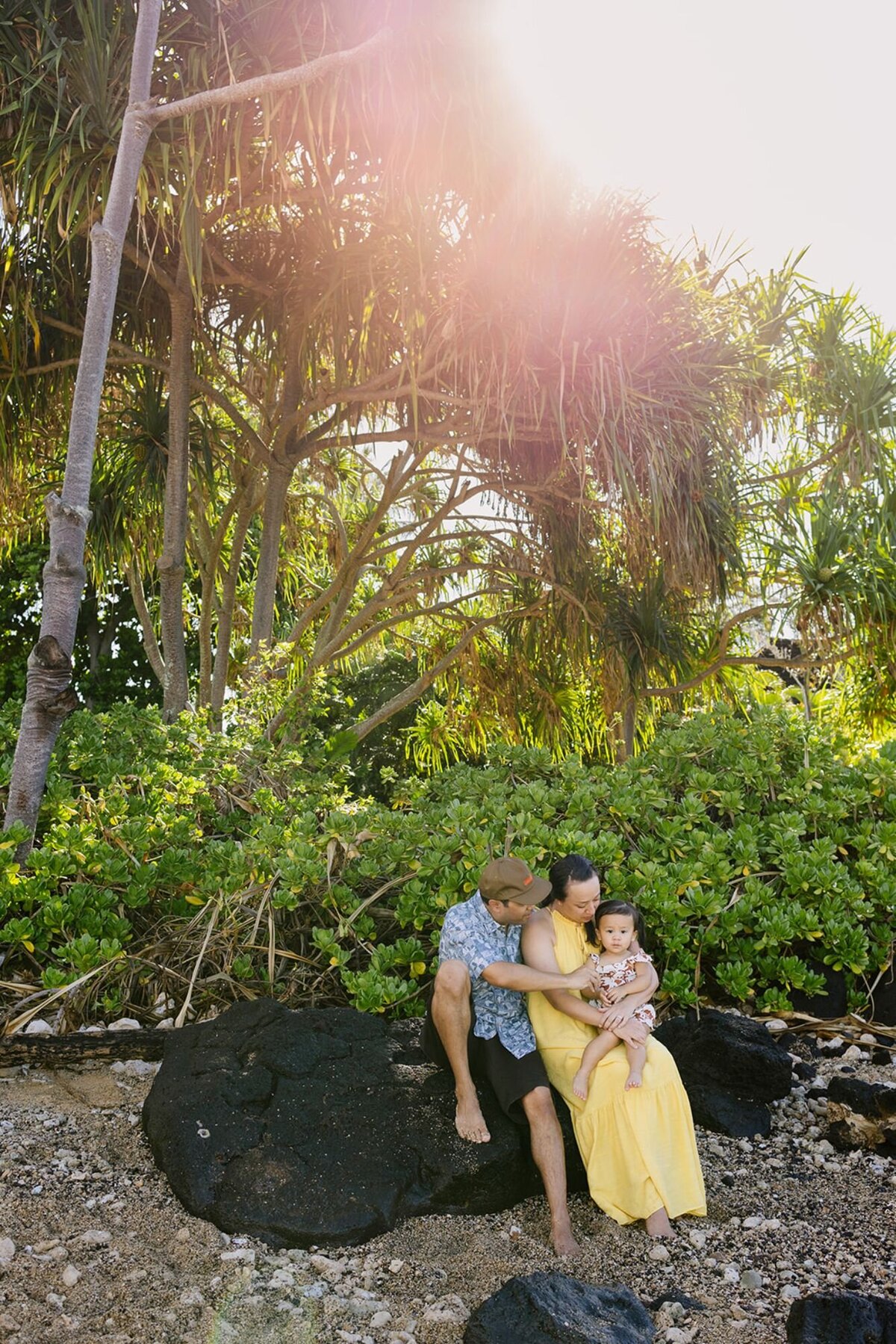A husband and wife hold their baby on a large lava rock on the beach of Kauai.
