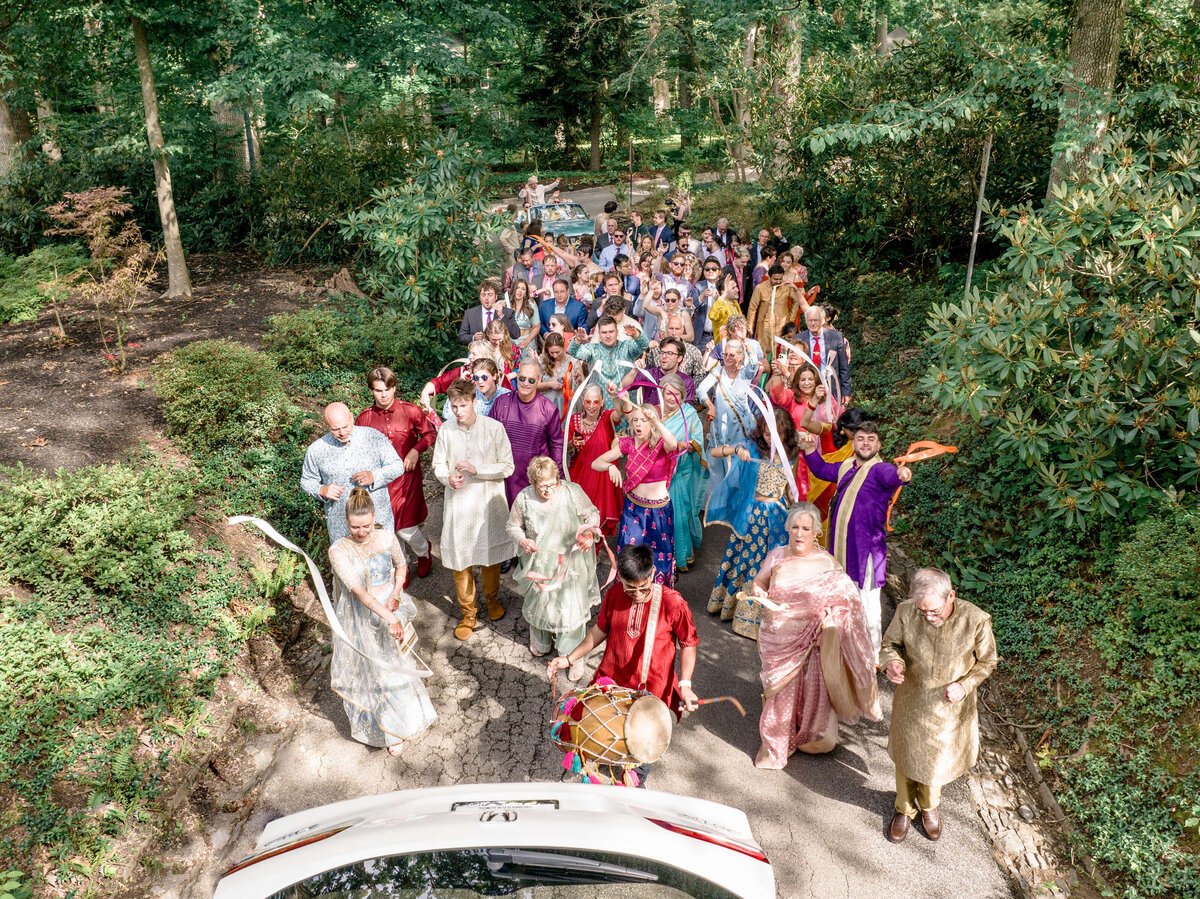 A vibrant group of people dressed in colorful traditional attire parade joyfully along a garden path. Some carry drums, others wave scarves, and greenery surrounds them, creating a festive atmosphere.