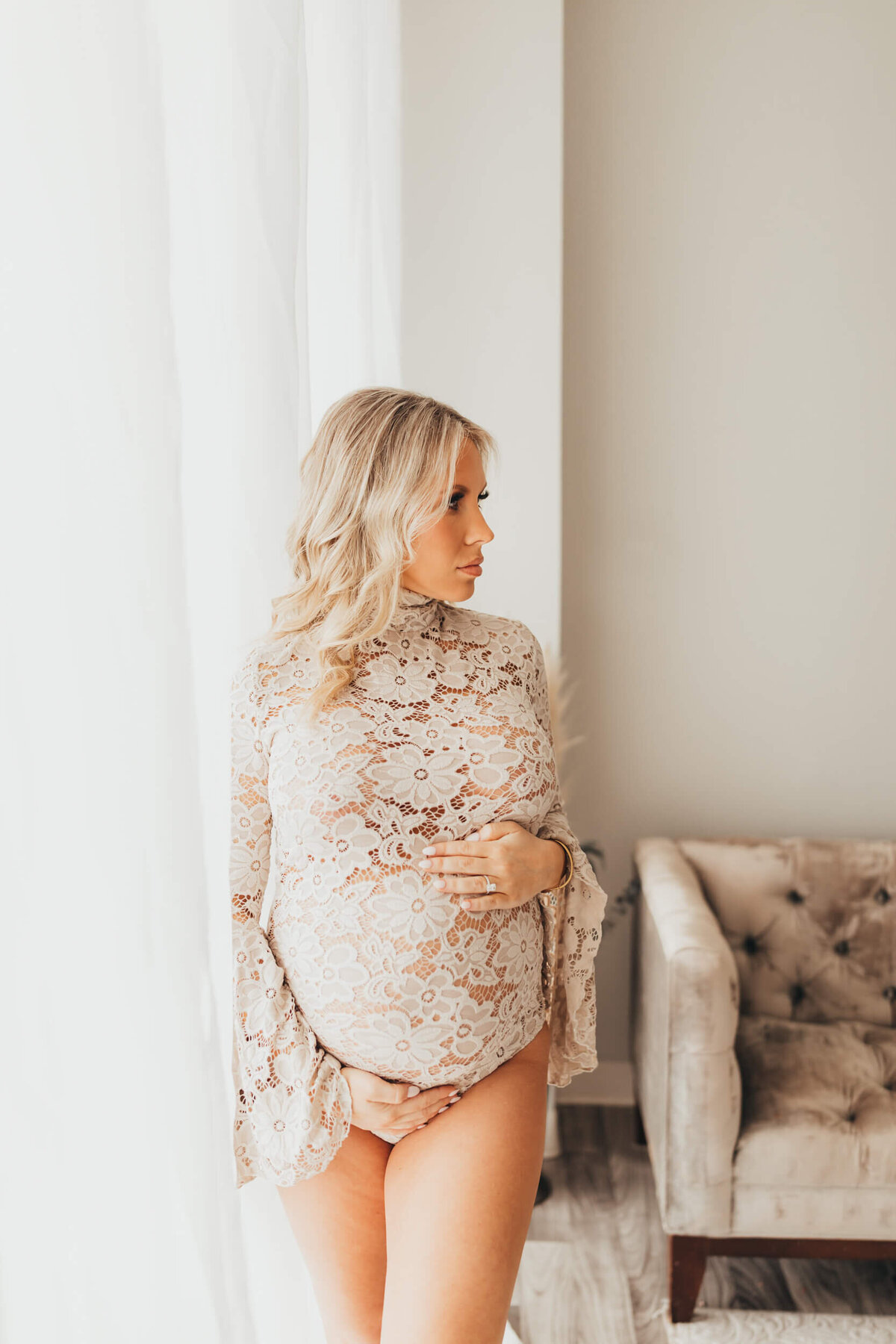 woman looks away, deep in thought, about her baby boy being born soon while standing in front of window, wearing nude bodysuit.