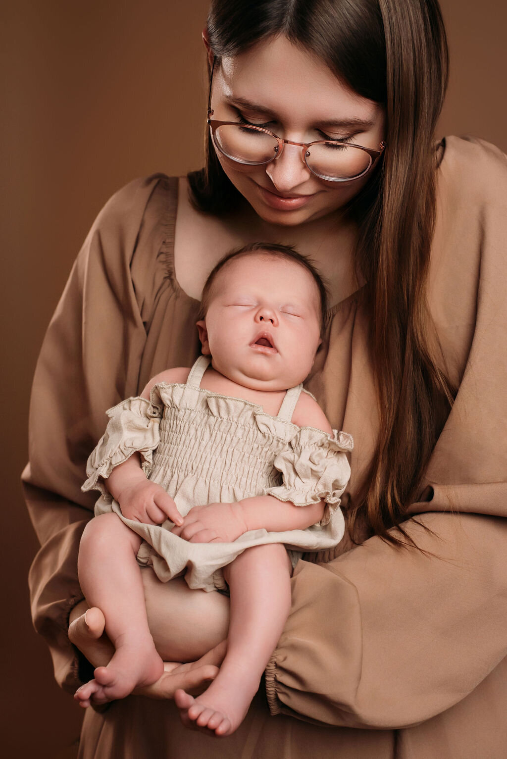 A woman in a tan boho dress sits in front of a neutral-toned backdrop holding a newborn baby in a beige outfit, captured by a photographer in Bemidji, Minnesota.
