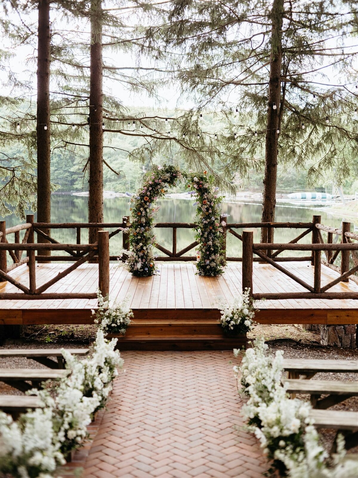 Stunning floral arbor by Canvas Weddings sits on wooden dock overlooking lake and trees at Cedar Lakes Estate wedding venue in Catskills; aisle lined with white florals