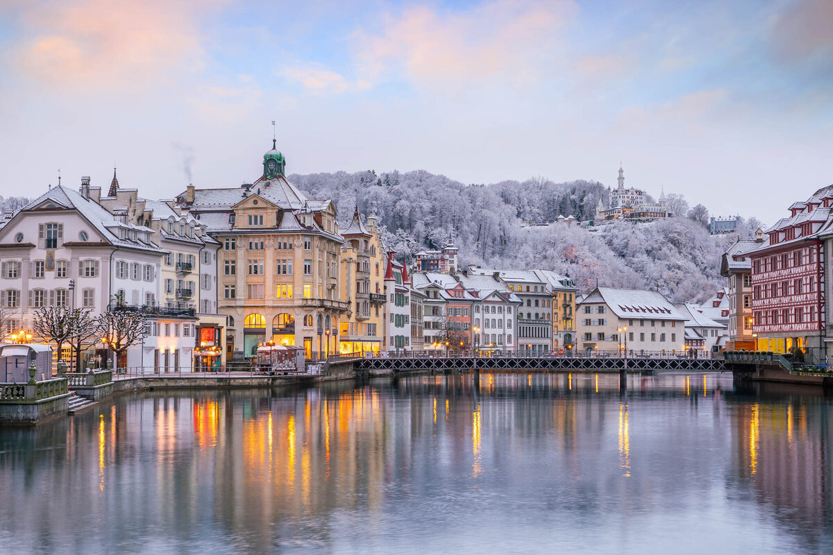 Scenic view of a European town in winter, with pastel-colored historic buildings reflected in a calm river. A snow-covered hill with trees and a distant structure rises in the background under a cloudy sky.