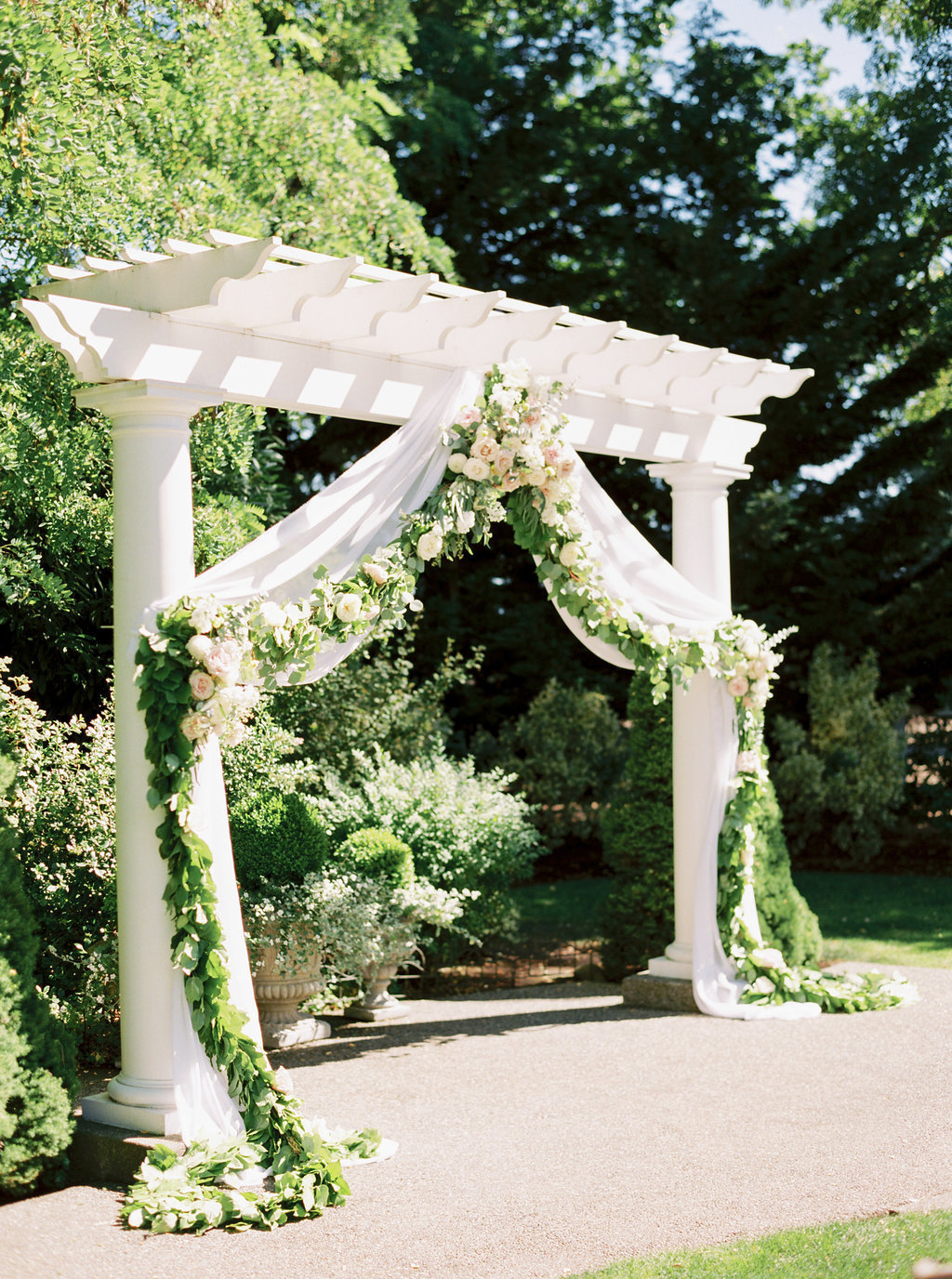 Beautiful outdoor garden wedding ceremony, white pergola draped with a greenery garland and floral.