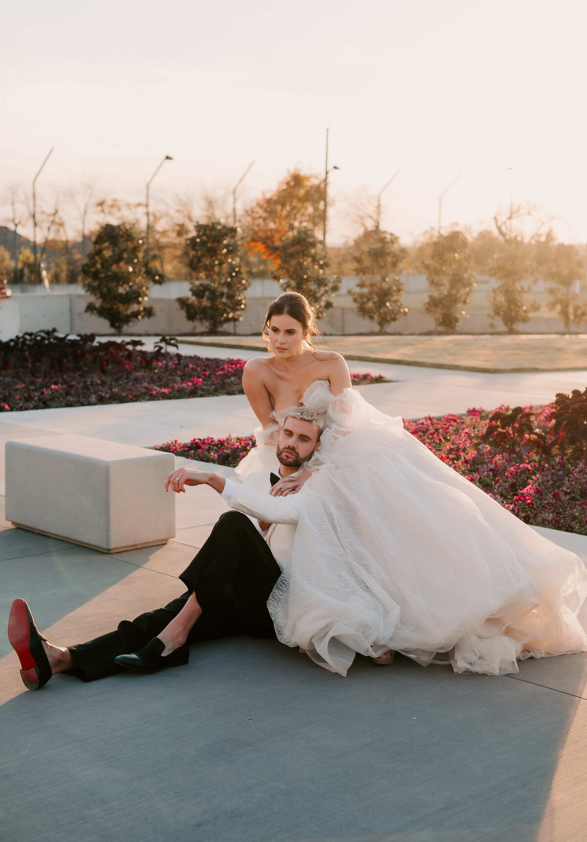 Bride in a flowing white gown sits on the ground beside the groom reclining on her lap. They are outdoors, surrounded by flowers and sunset light. Romantic and serene.