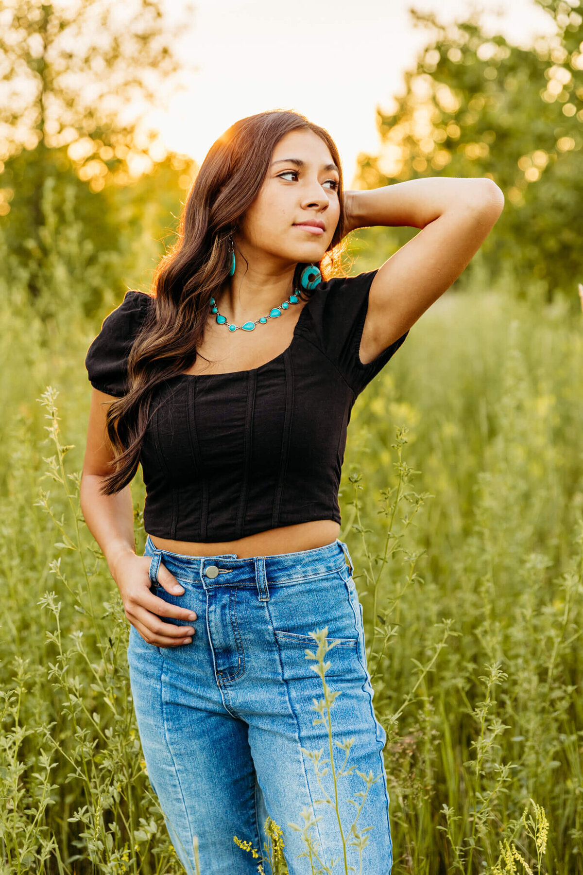 senior girl standing in tall grass with one hand in hair and other in belt loop as she looks over shoulder and the sun sets behind her captured by Ashley Kalbus Photography