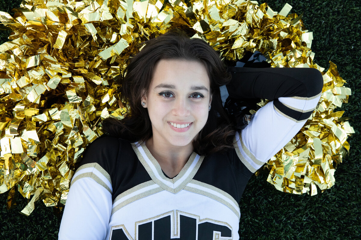A teenage girl in a black and white cheerleading uniform lays on gold pom poms Nanuet senior portrait photographer Milestone Images