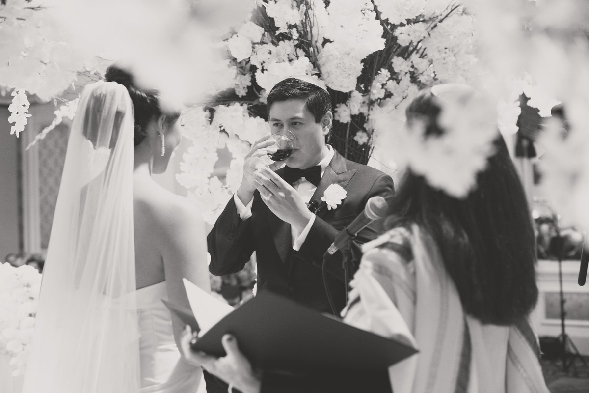 black and white photo of groom drinking from the glass of wine during wine ceremony for wedding at The Garden City Hotel