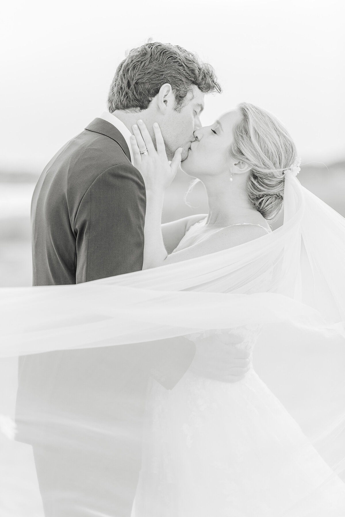 A bride and groom share a kiss along the shores of the Wychmere Beach Club in Harwichport, MA for a formal wedding portrait. The bride's hands are gently resting on her groom's neck, the groom's hands resting around the bride's waist. The bride's veil is blowing in front of them. Captured by MA Wedding Photographer Lia Rose Weddings