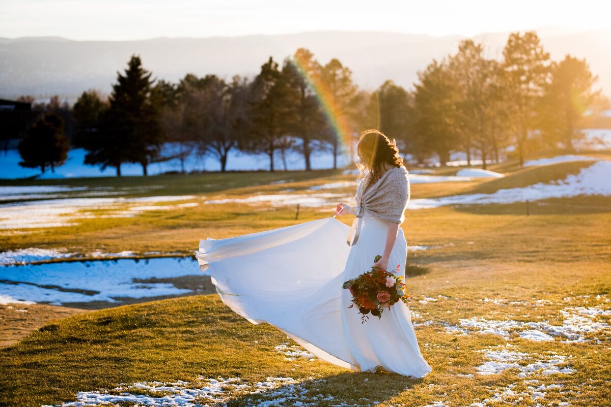 A bride twirls her dress while standing in a field with snow in the background and a rainbow reflecting off the camera lens.