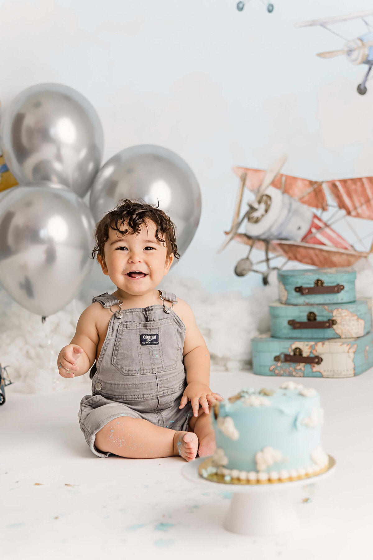 baby boy in grey overalls smiling with smashed cake on his legs and face