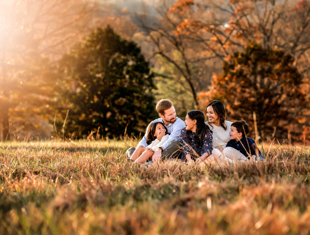 A family of five sitting in the grass and laughing with trees behind them