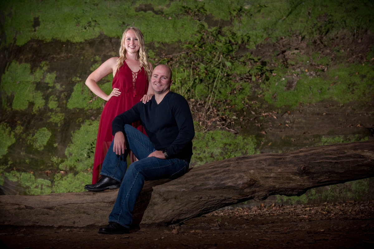 Red dressed bride and her groom pose on a log against a green moss covered wall  of a waterfall at staved rock state park