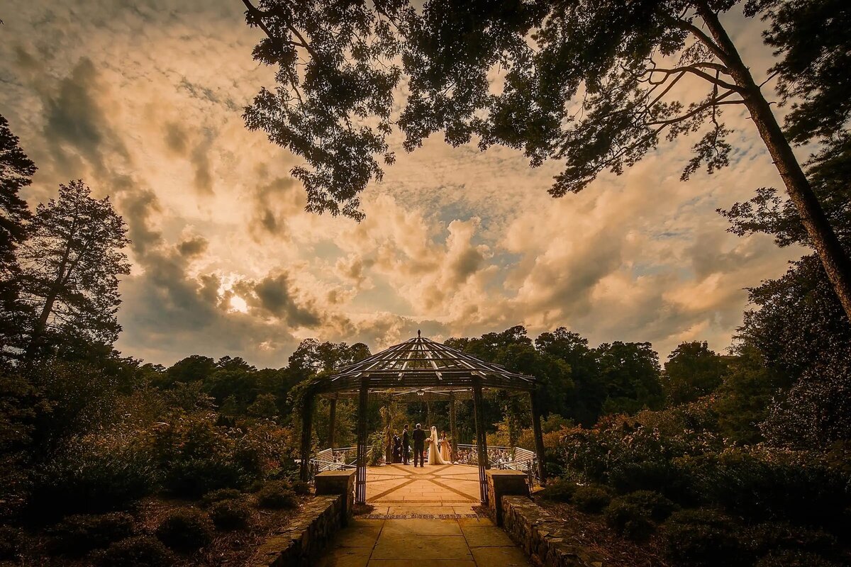 A stunning sunset provides a dramatic backdrop for an outdoor wedding ceremony framed by an ornate gazebo