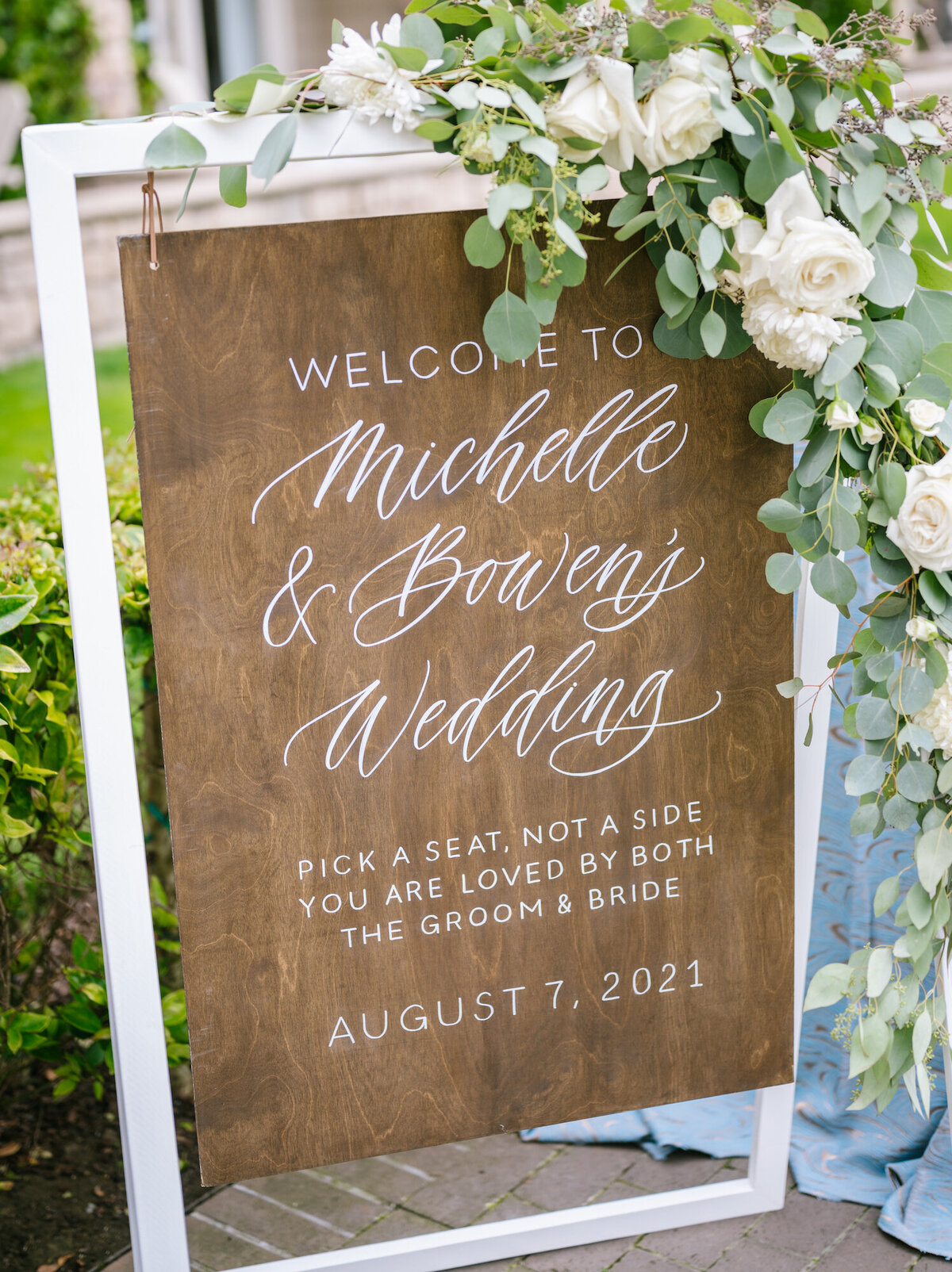 Wood welcome and ceremony aisle sign for a wedding, decorated with florals