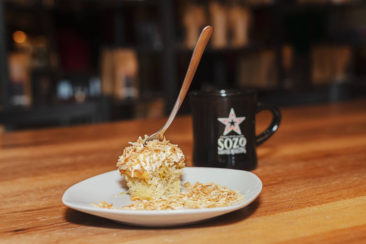 A slice of coconut-topped cake on a white plate with a wooden spoon, next to a black coffee mug with a star logo, on a wooden table.