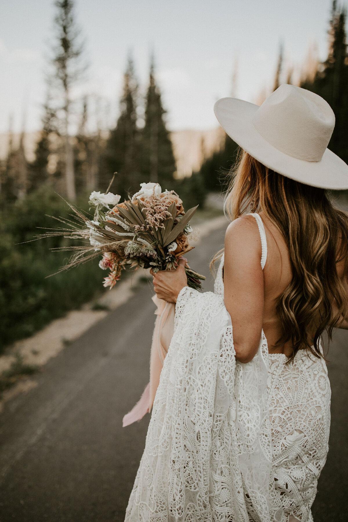 A bride wearing a white wedding gown and ivory hat holding a bouquet of blush and white flowers overlooking  mountains.