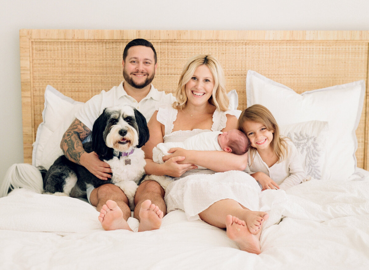 Family sits on a bed with their newborn and dog during a newborn session in Raleigh NC. Photographed by Raleigh Newborn Photographer A.J. Dunlap Photography.
