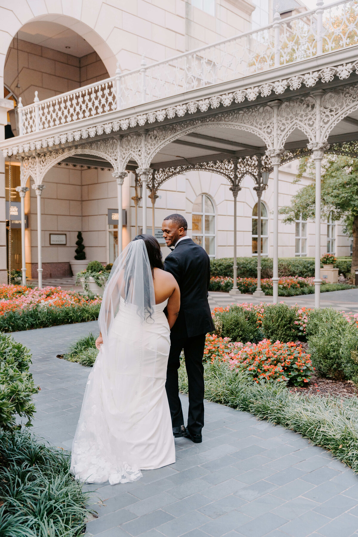 A bride in a white gown and veil walks arm-in-arm with a groom in a black suit along a garden path, surrounded by flowers and ornate architecture.
