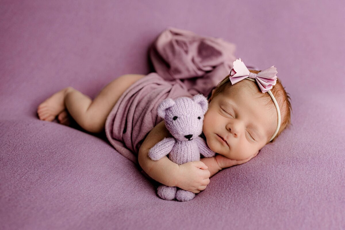 newborn baby girl is posed laying on her side cuddling a  purple bear wrapped in a purple wrap and wearing a matching bow
