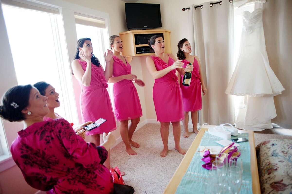 bridesmaids in colorful pink dresses look in surprise as they pop the cork from champagne