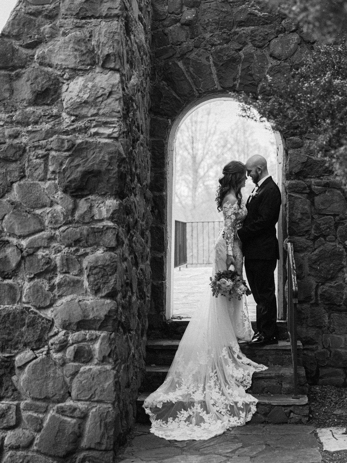 A bride and groom stand together foreheads touching under a stone archway as the bides's lace dress train falls elegantly down a set of stairs