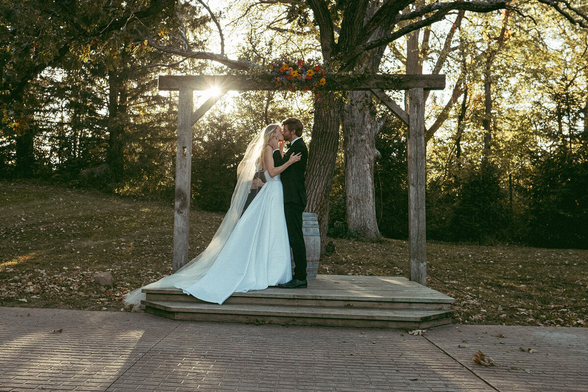 A bride and groom sharing a kiss under a wooden archway adorned with flowers in a park setting at sunset during their Iowa wedding.