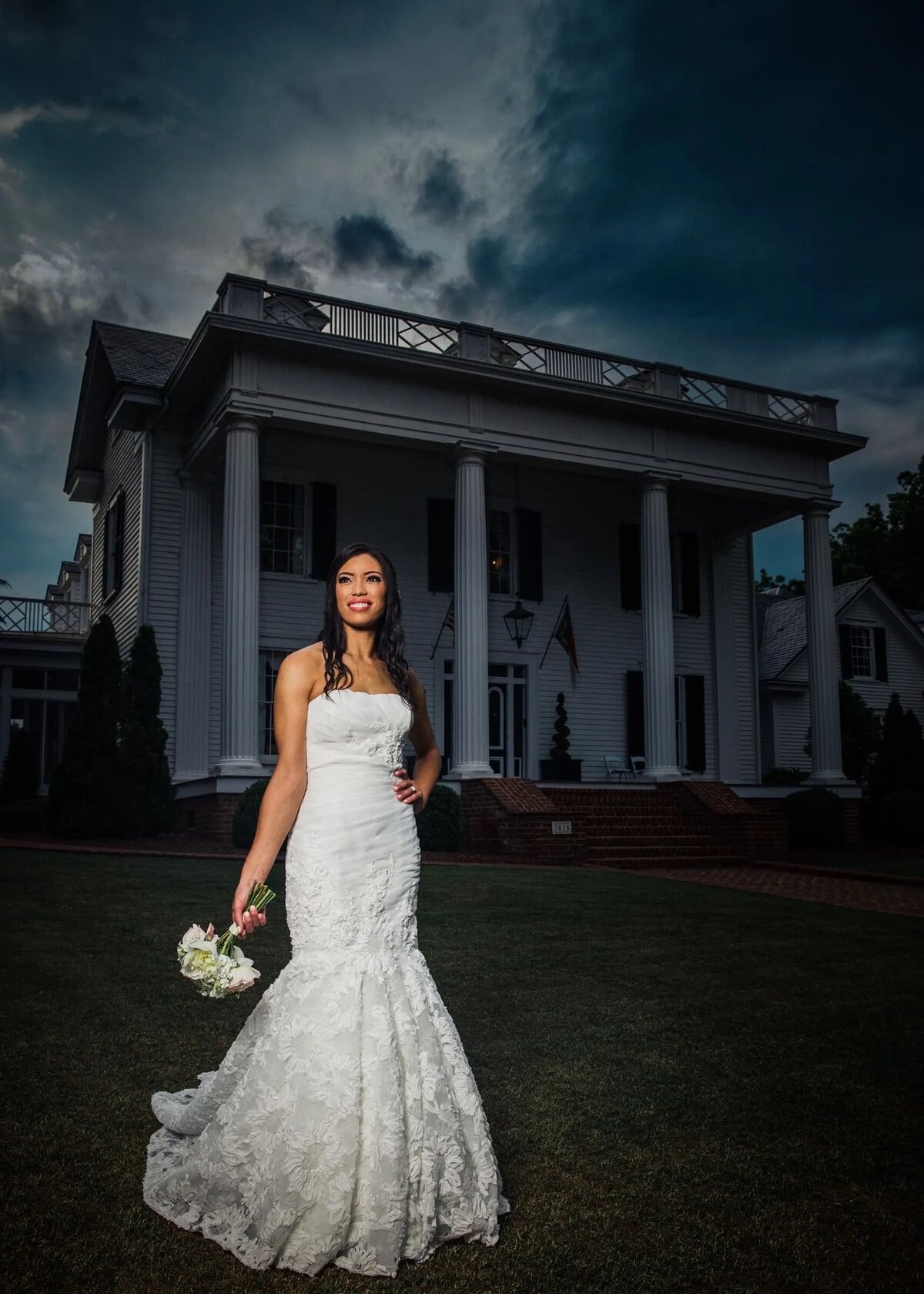 A bride standing in front of a large house with one hand on her hip.