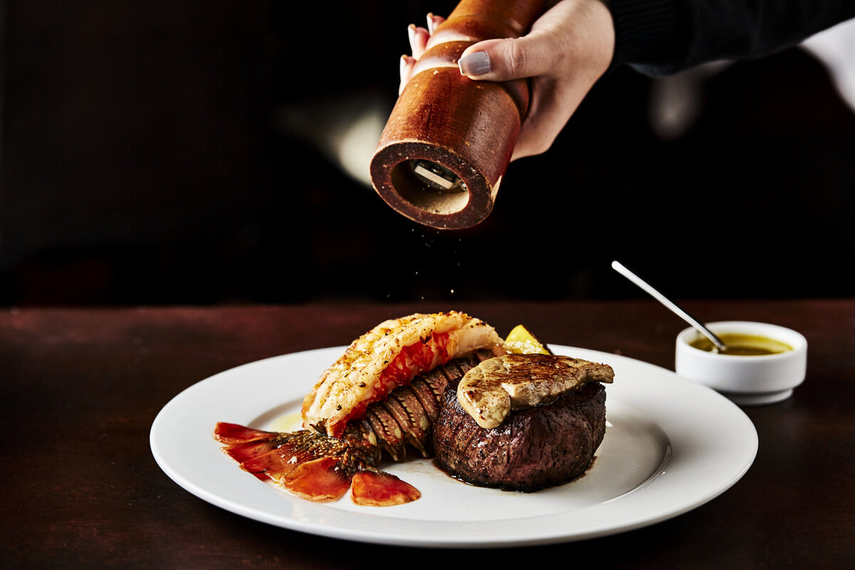 Pepper being cracked on top of a plate with lobster and steak.