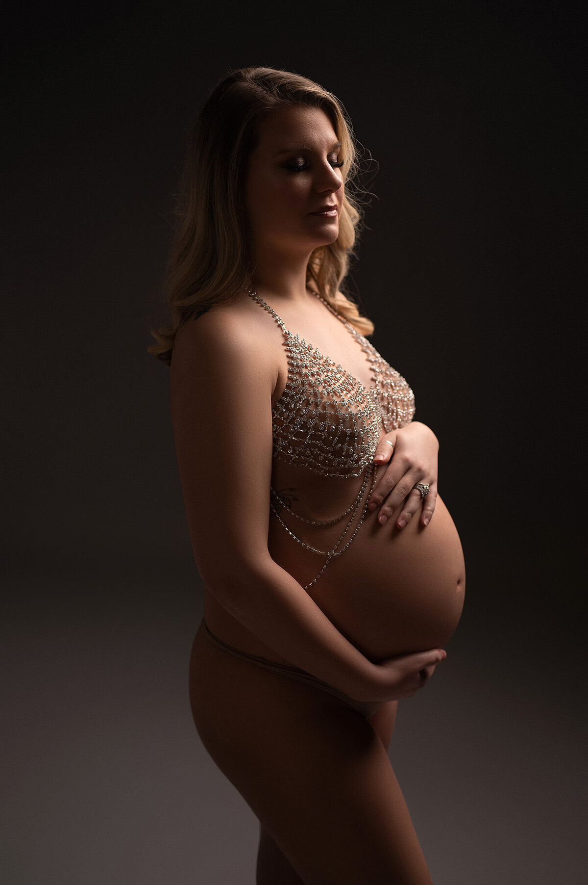 Portrait of a pregnant woman cradleing her belly while wearing beaded lingerie in front of a gray backdrop in our Waukesha studio.