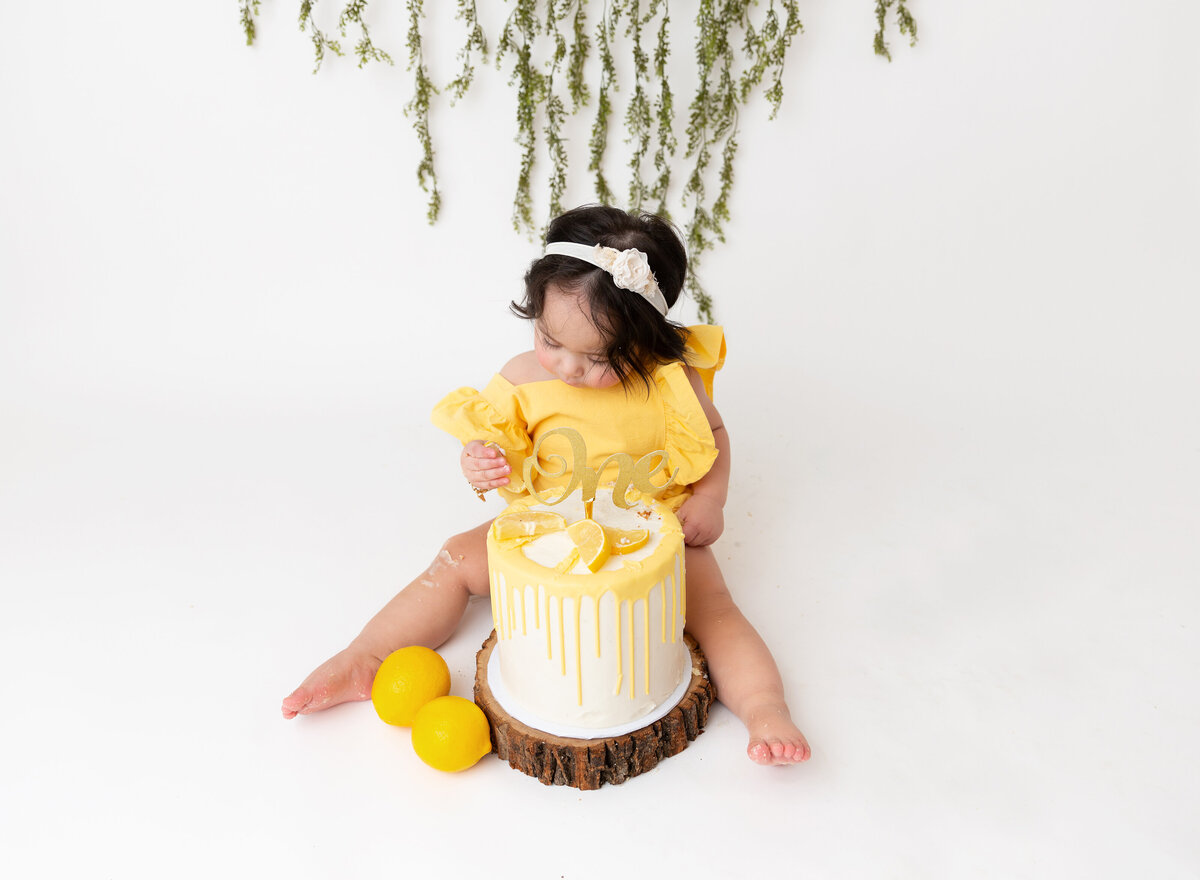 Baby girl sits with lemon-themed cake for her first birthday cake smash. She is wearing a yellow romper and looking at the icing on her fingers.
