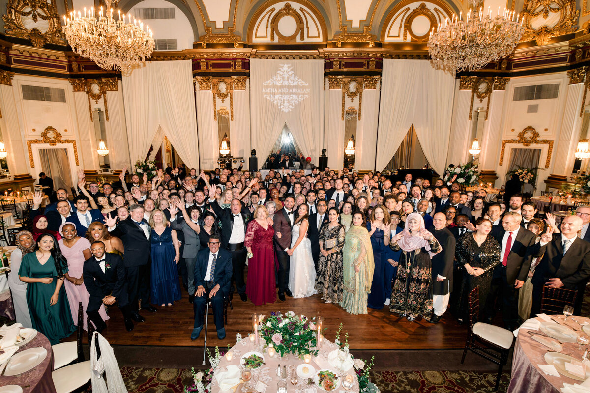 A large group of people dressed in formal attire gather in an opulent ballroom with ornate chandeliers and draped walls. They are smiling and posing for a group photo in front of elegantly set tables.