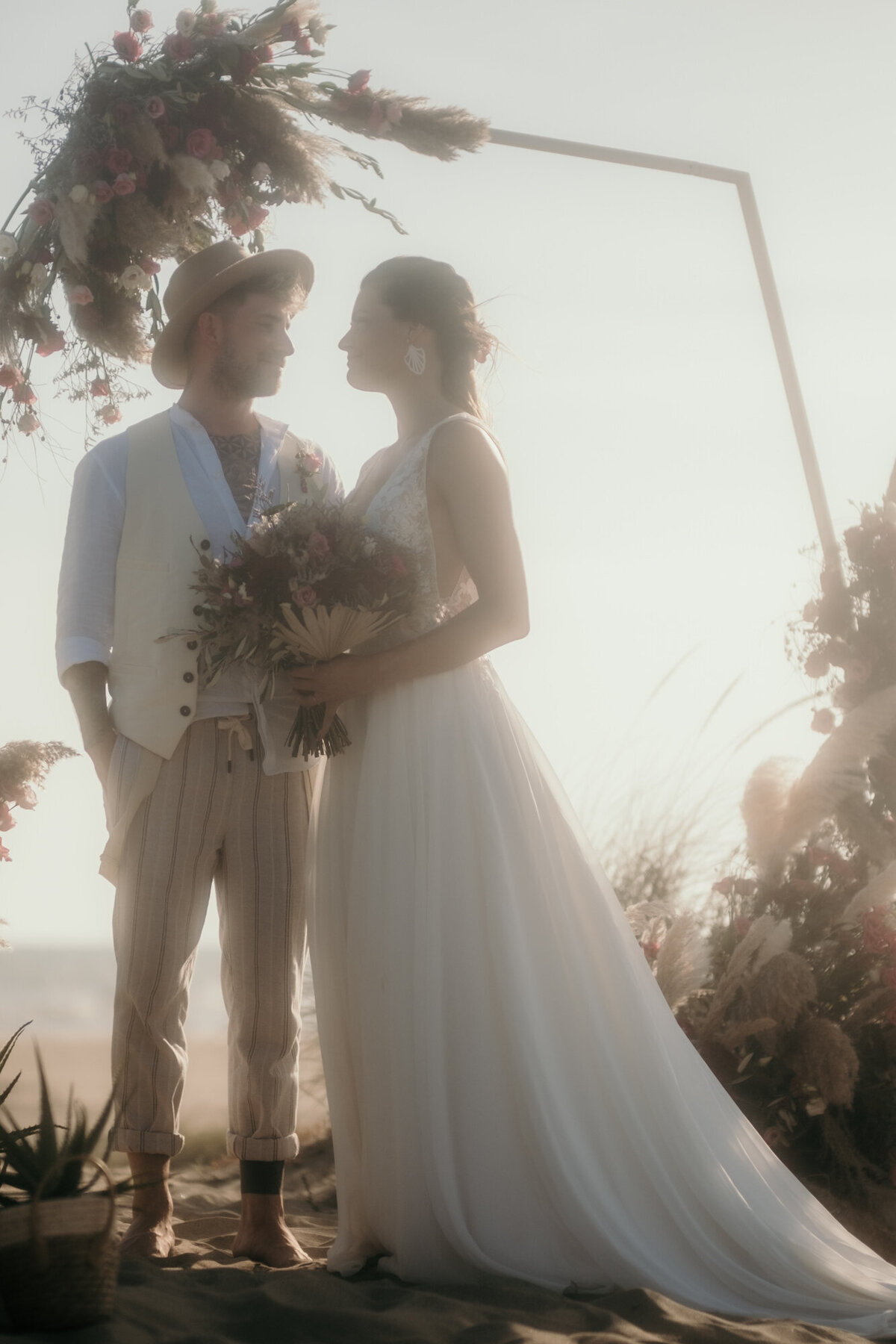 Die Sonne strahlt von hinten auf das sich glücklich anblickende, unter dem Hochzeitsbogen stehende Paar.