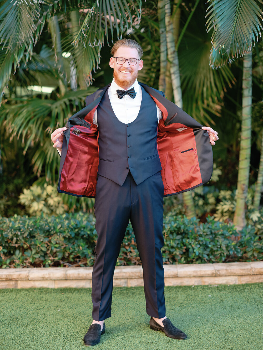 Handsome groom in a navy blue tuxedo with red satin lining and an emerald green bowtie, captured in a portrait at the Don Cesar Hotel in St. Pete Beach, FL, during a December wedding.
