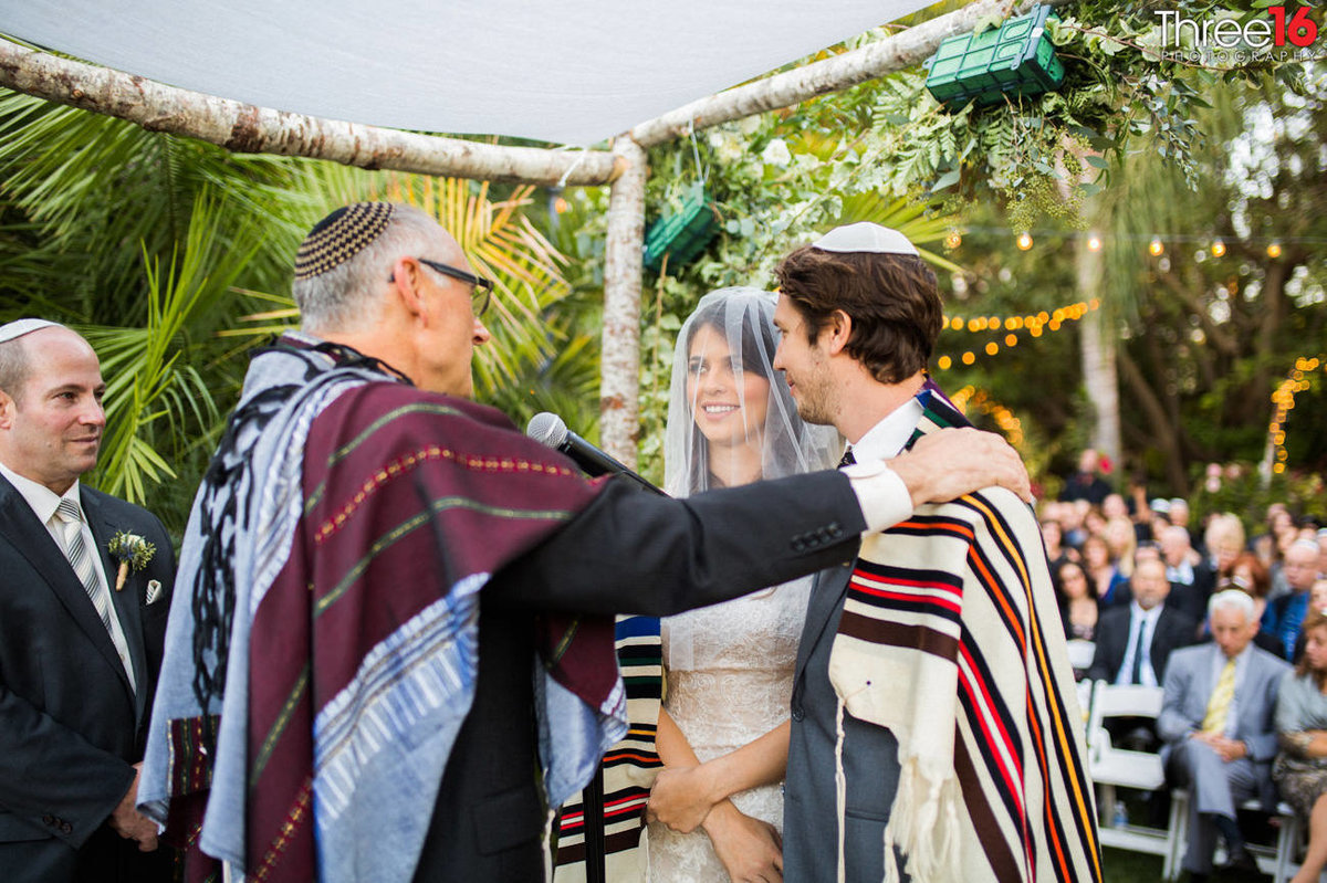 Rabbi places his hand on the Groom's shoulder as he speaks to him and the Bride during the wedding ceremony