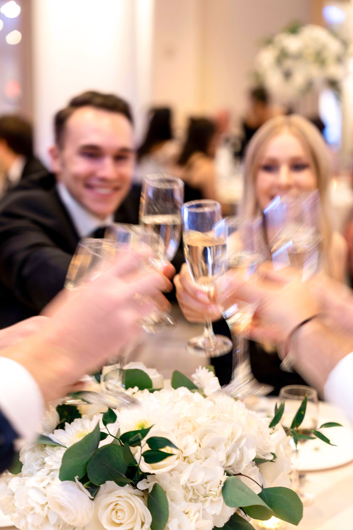 A group of people raising champagne glasses in a toast at a celebration. They are smiling, with a floral arrangement of white roses and greenery on the table in the foreground. The background is softly blurred, highlighting the festive atmosphere.