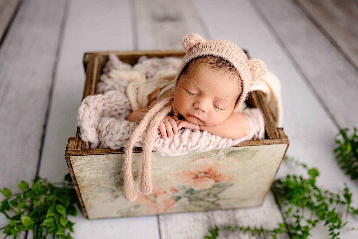 newbonr baby girl is posed in a crate with flower decoration wearing a peach bonnet
