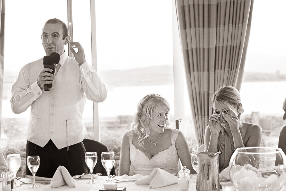 Groom in white waistcoat giving a speech at the top table in the Dingle Skellig Hotel while the bride laughs and the bridesmaid crys