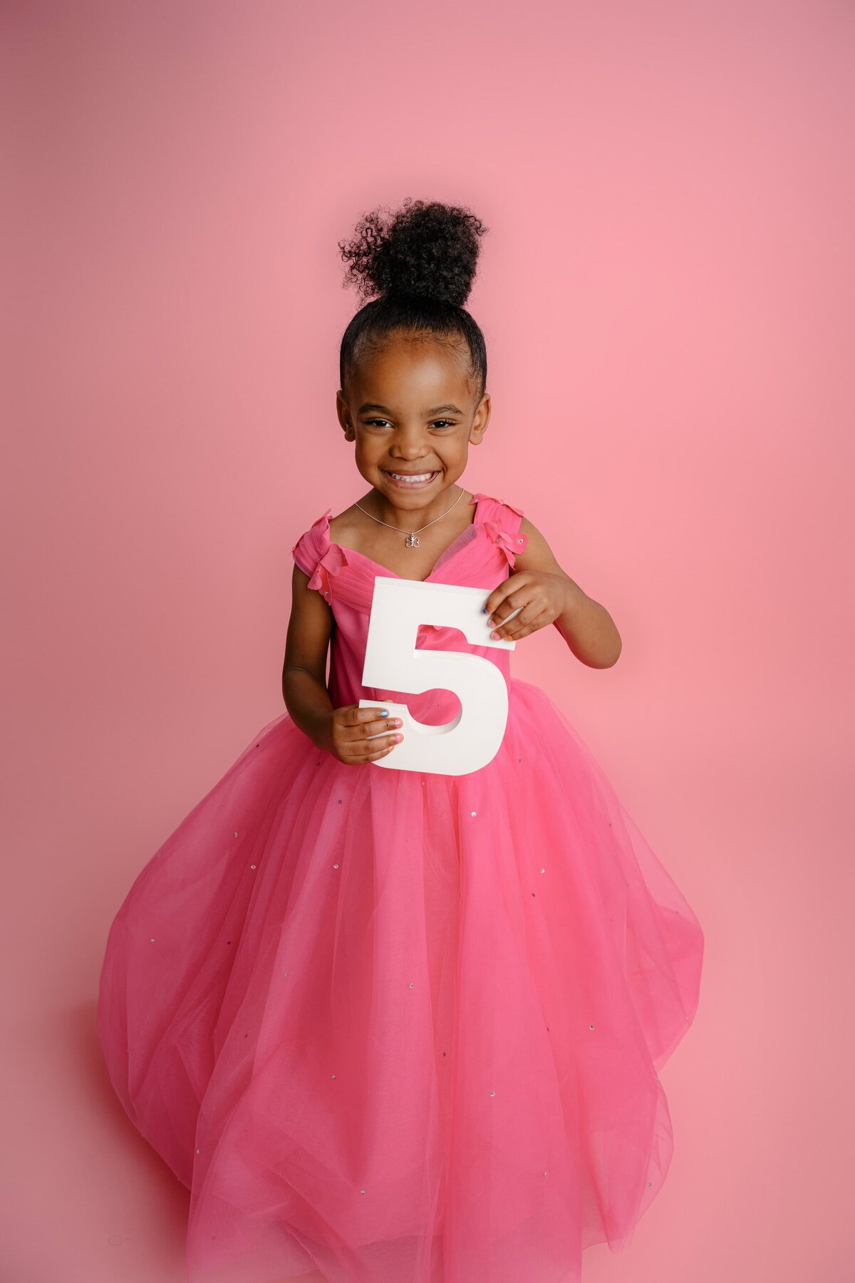 Little girl holds a number five prop while dressed in a pink dress with a twirl bottom on a pink background in a Charlotte studio location