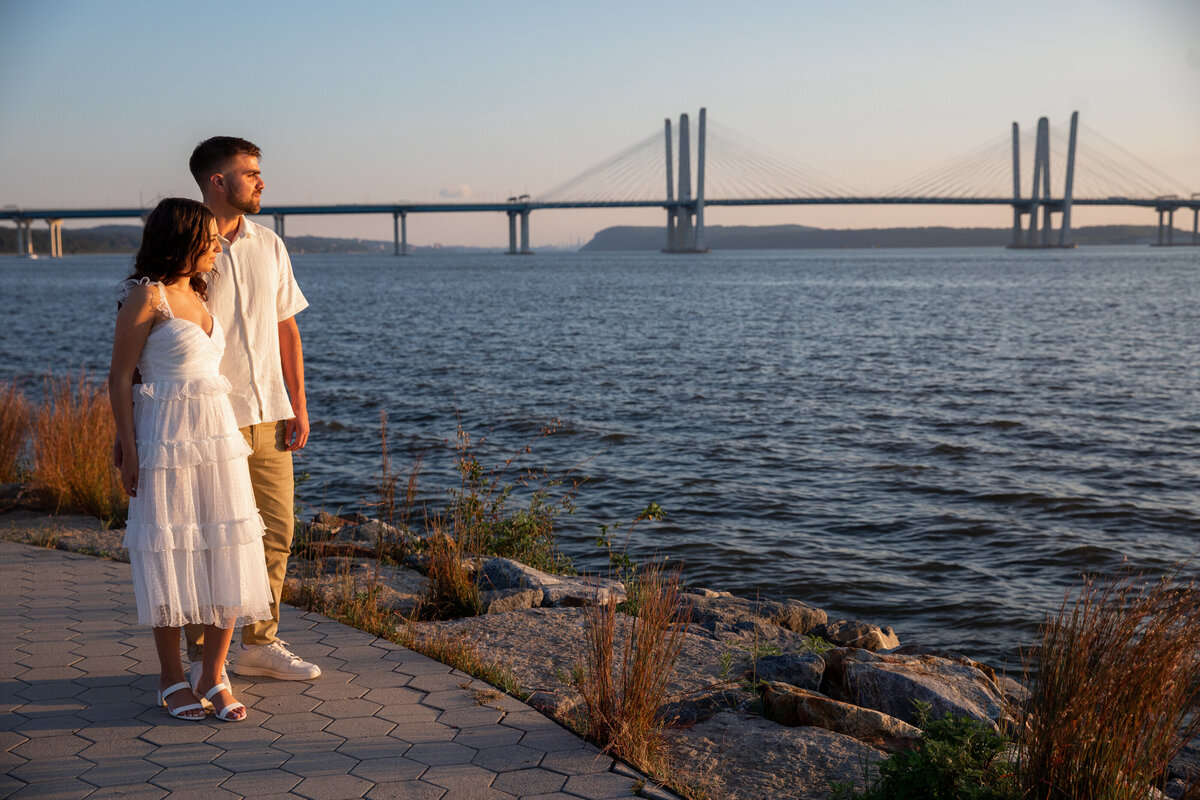 A white could in white clothing stand on the banks of the Hudson River with the Mario Cuomo Tappan Zee bridge in the background Westchester engagement session New York wedding photographer