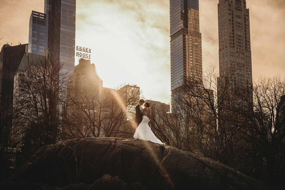 bride and groom stand on rock on sunlight shines on them with skyscrapers in background photo by cair fletcher photography