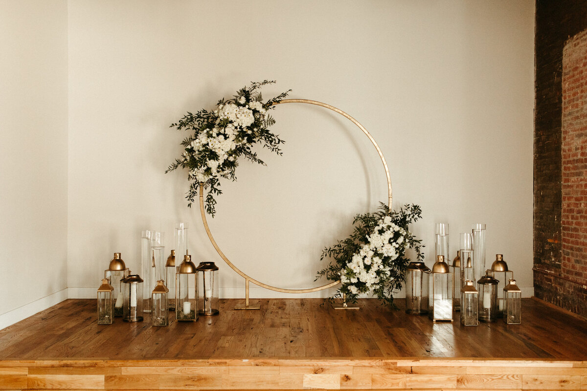 Round hoop arch with floral decor against white wall with candles inside lanterns sitting around the base on a wooden floor